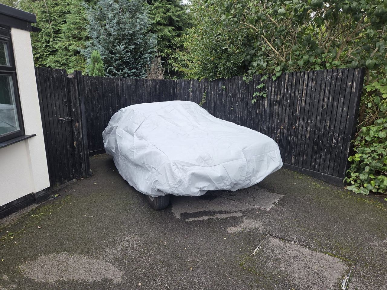 A light grey car cover fitted over Bob in a paved yard, against a black fence, with one tyre still showing.
