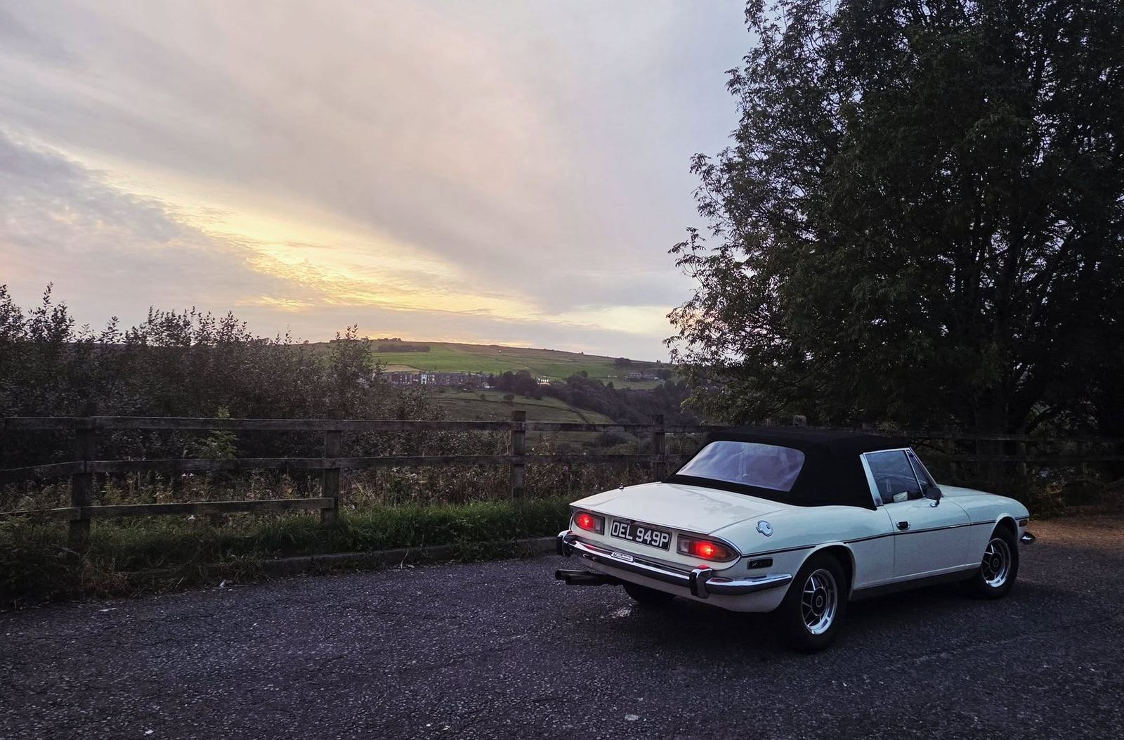 White 1975 Triumph Stag Bob, black soft top, parked at dusk beside a wooden fence with hills behind.
