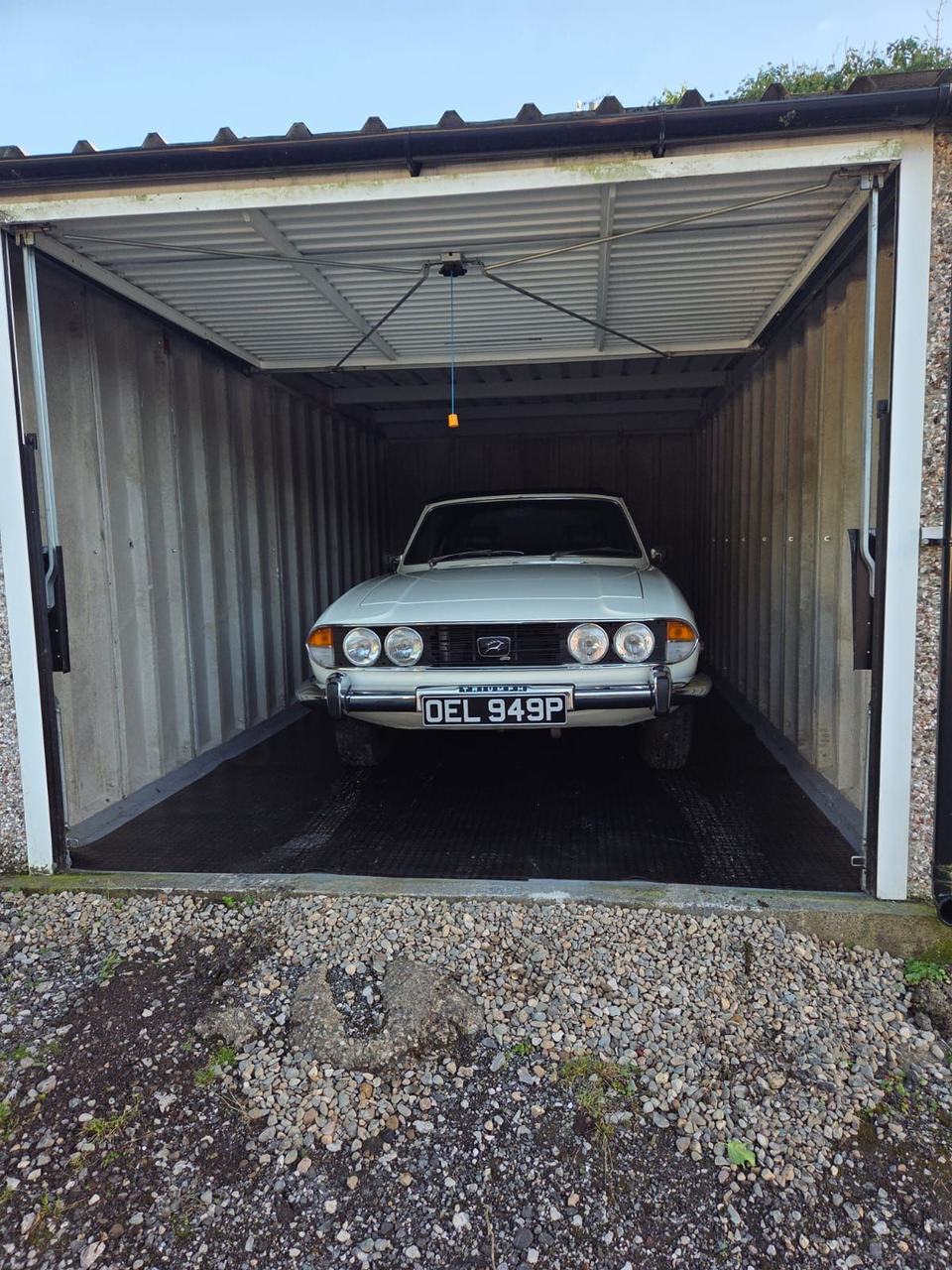 Triumph White Stag, registration OEL 949P, parked nose-out on black rubber matting inside a corrugated metal garage.